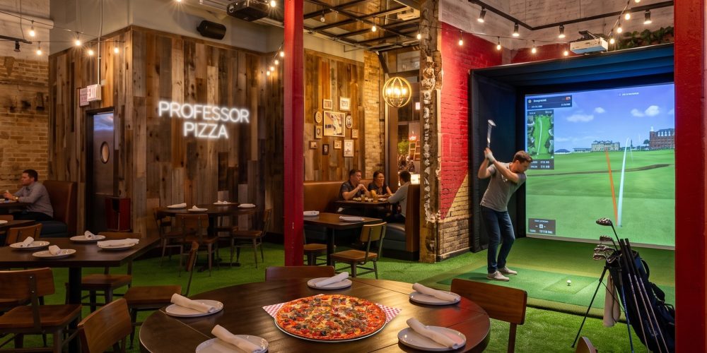 Golfer taking a swing in a pro-grade golf simulator bay at Professor Pizza, surrounded by green turf flooring and fresh Professor Pizza in Old Town Chicago.